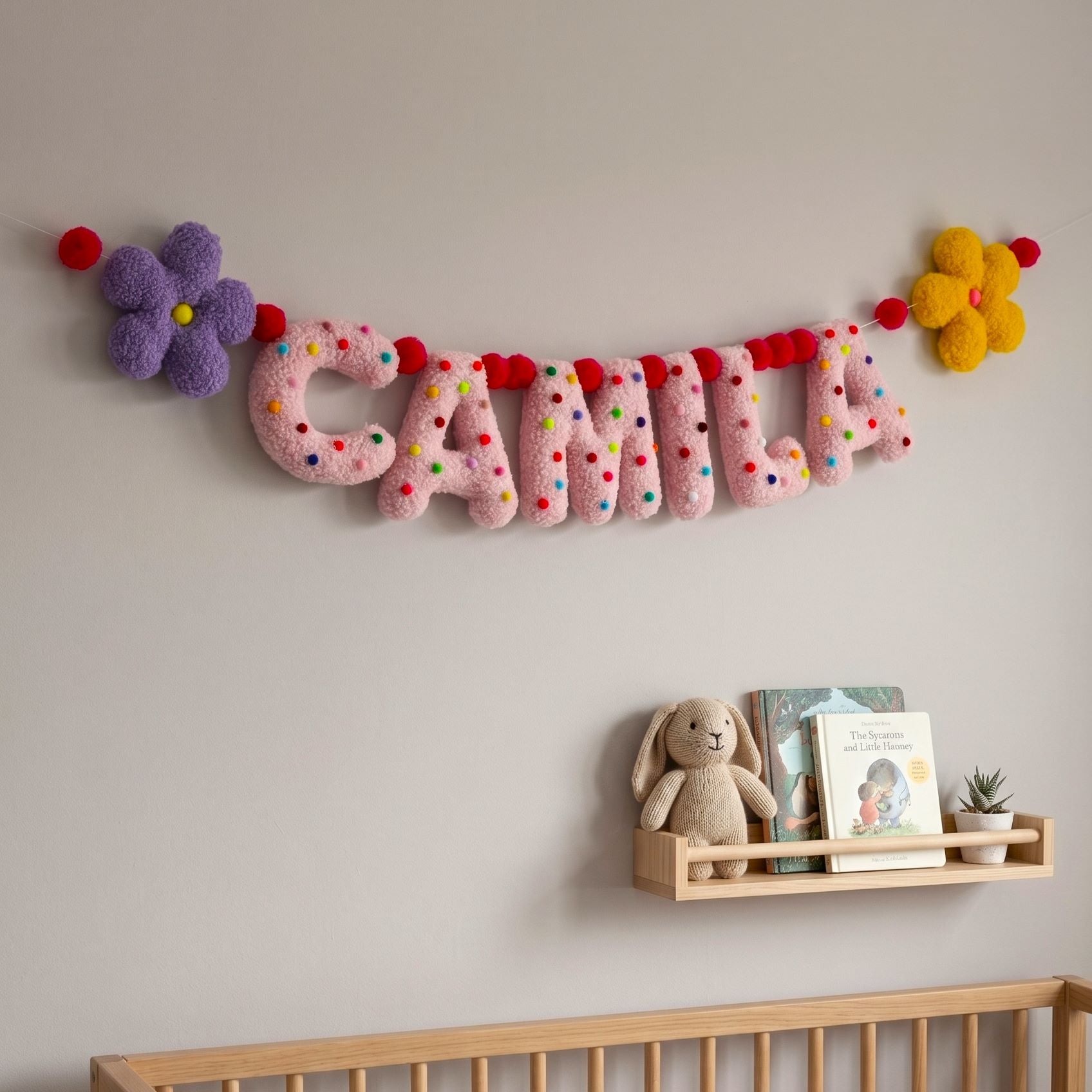 Decorative name banner with colorful letters on a wall above a shelf with a teddy bear and books.