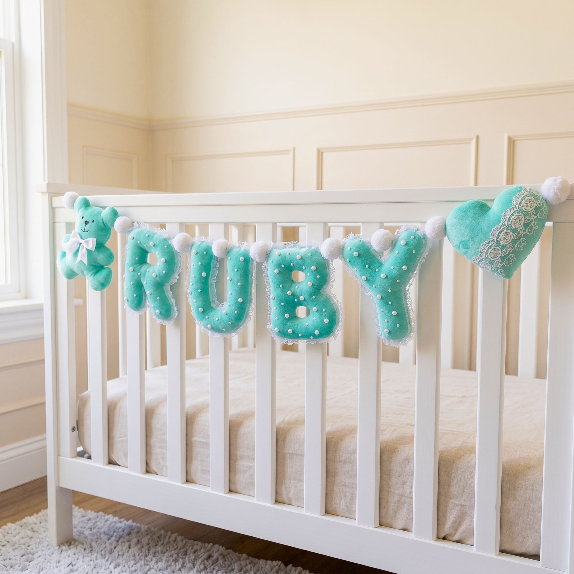 A white wooden baby crib with a plush letter banner spelling 'RUBY' in mint green, accompanied by a small plush bear and a heart-shaped ornament, set in a softly lit nursery.
