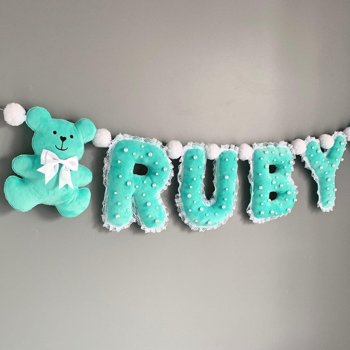 A personalized baby nursery wall banner spelling 'RUBY' in mint green letters with white pom-pom trim, accompanied by a matching mint green teddy bear plush with a white bow, hung against a plain gray wall.