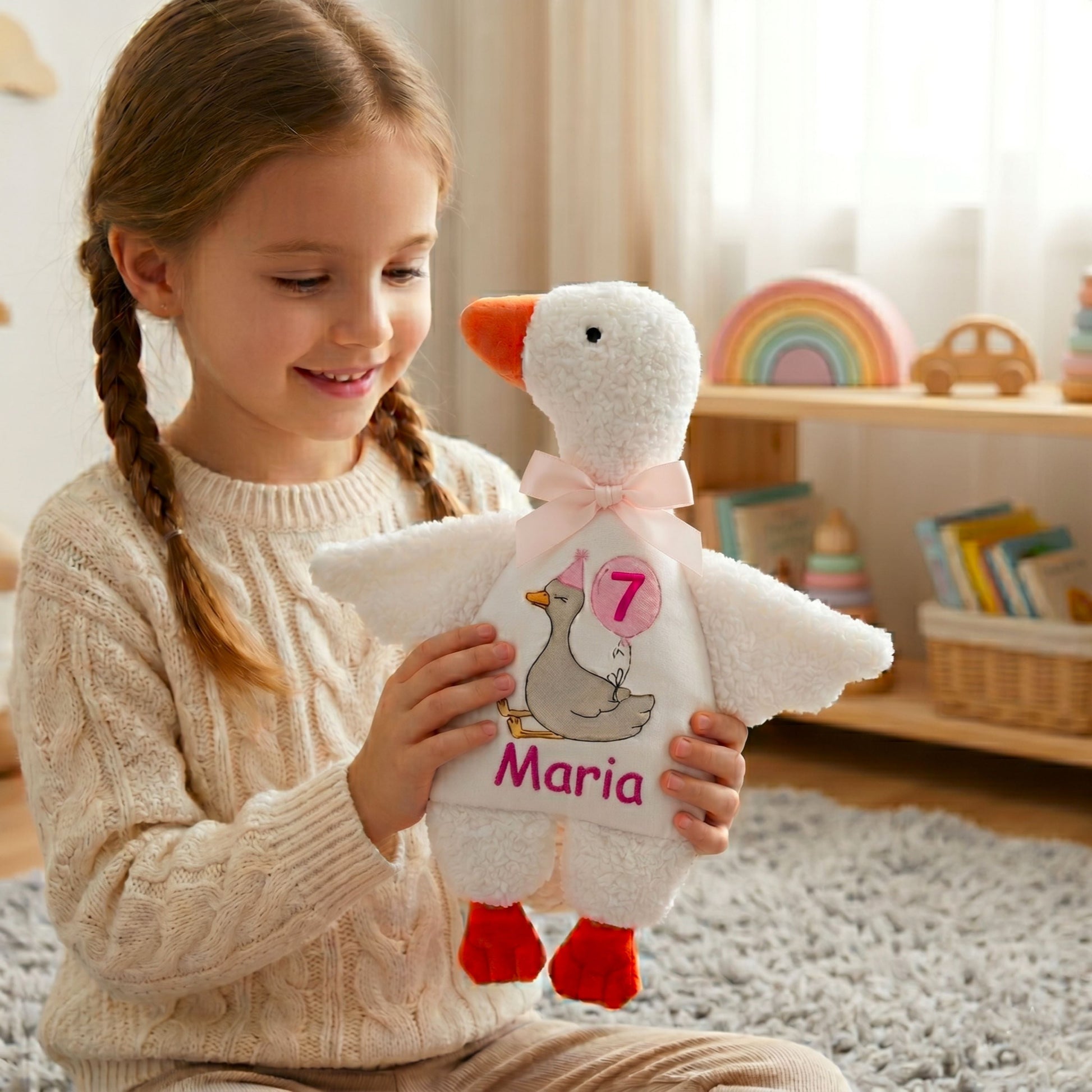 Happy little girl holding her personalized birthday plush goose toy in a playroom. Custom embroidered soft stuffed animal with the name "Maria" and age 7, perfect for kids room decor and cuddling.