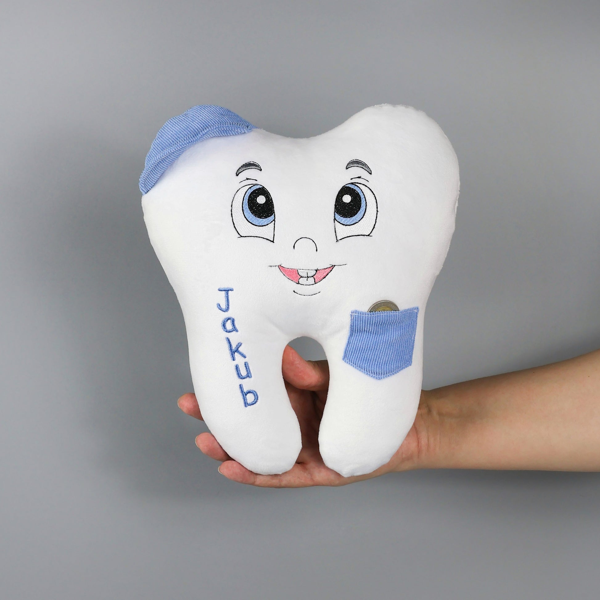 Plush tooth-shaped toy with cartoon face and blue accents, held by a hand against a gray background.