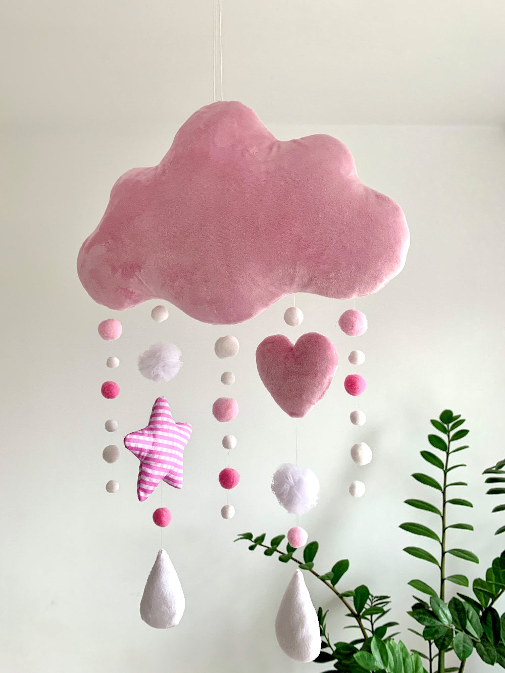 Pink cloud and heart-shaped decoration hanging against a white wall with green leaves at the bottom.