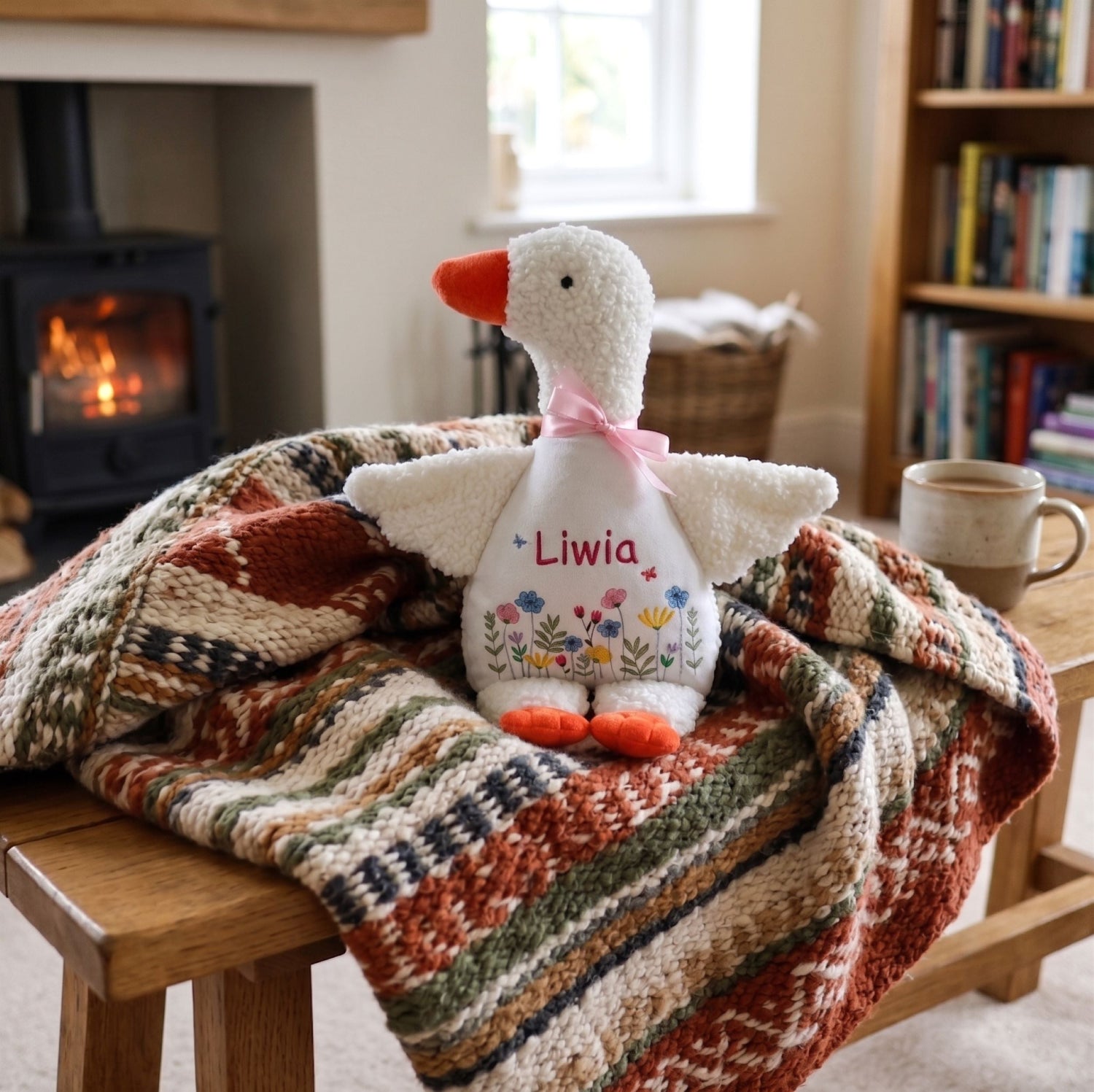 Stuffed goose toy with 'Liwia' embroidered on it, placed on a patterned blanket in a cozy room.