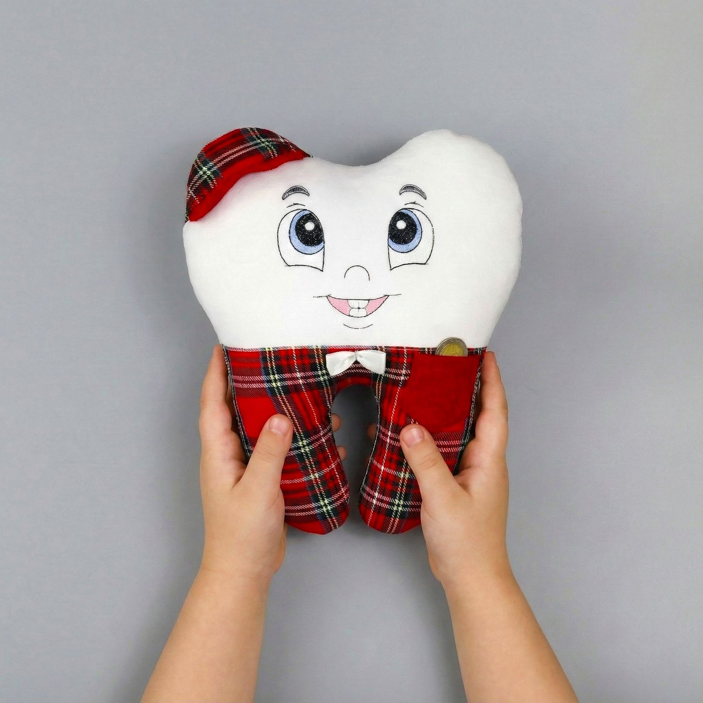 Plush toy tooth with plaid outfit held by hands against a gray background