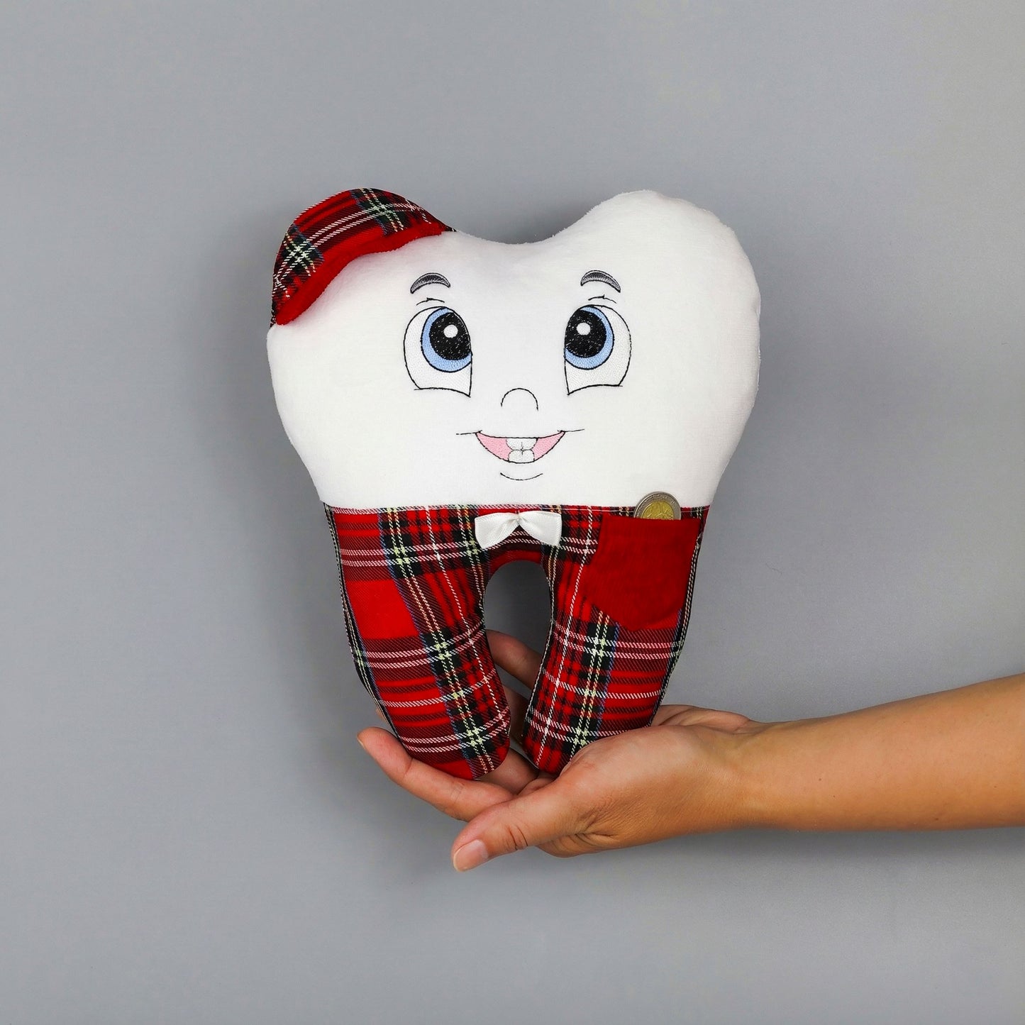 Plush tooth-shaped pillow with a face, plaid pants, and a bow tie held by a hand on a gray background