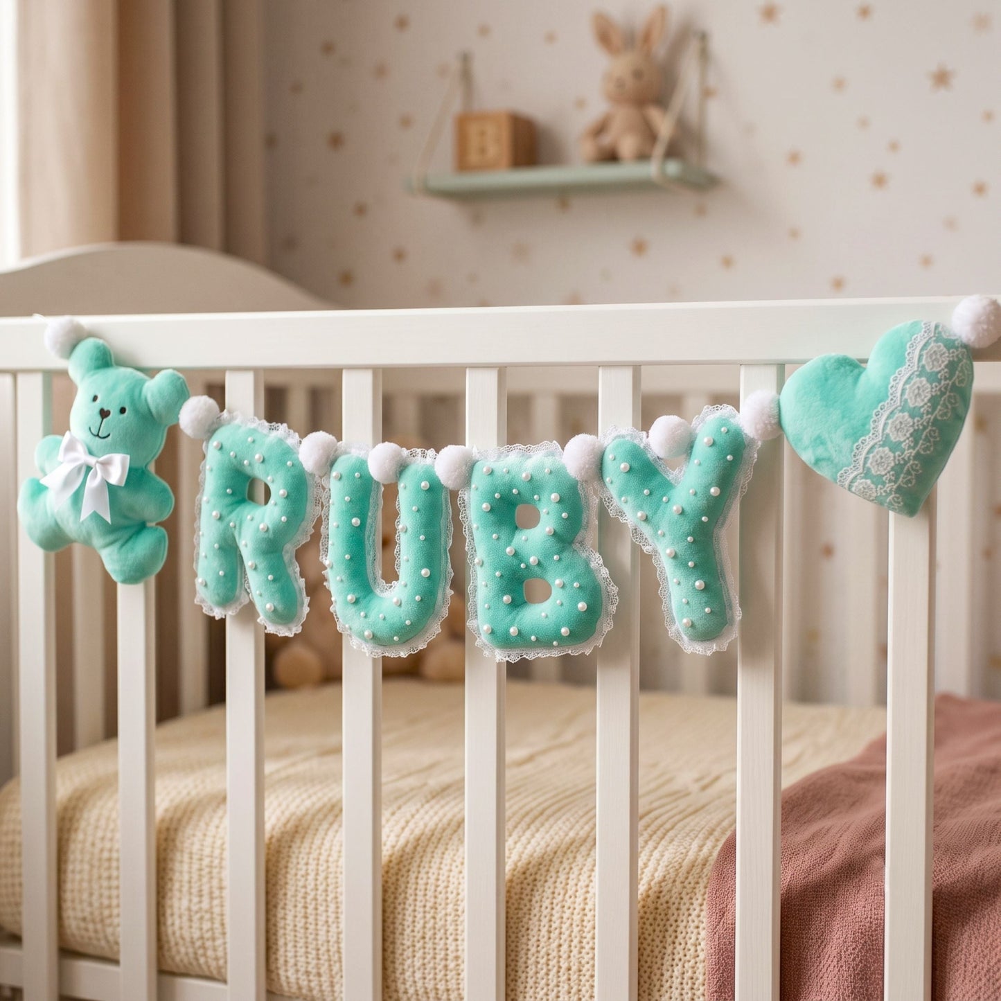 A baby nursery scene with a white crib, cream bedding, and a mint green felt letter banner spelling RUBY, plus a matching teddy bear and heart plush accents.