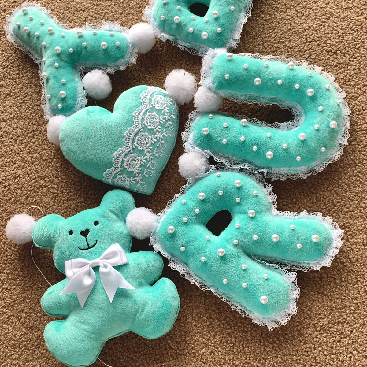 A set of handmade baby shower banner spelling out 'BABY' in mint green, featuring a teddy bear with a white bow, a lace-trimmed heart, and letter pillows decorated with white pearls, all arranged on a brown carpet.