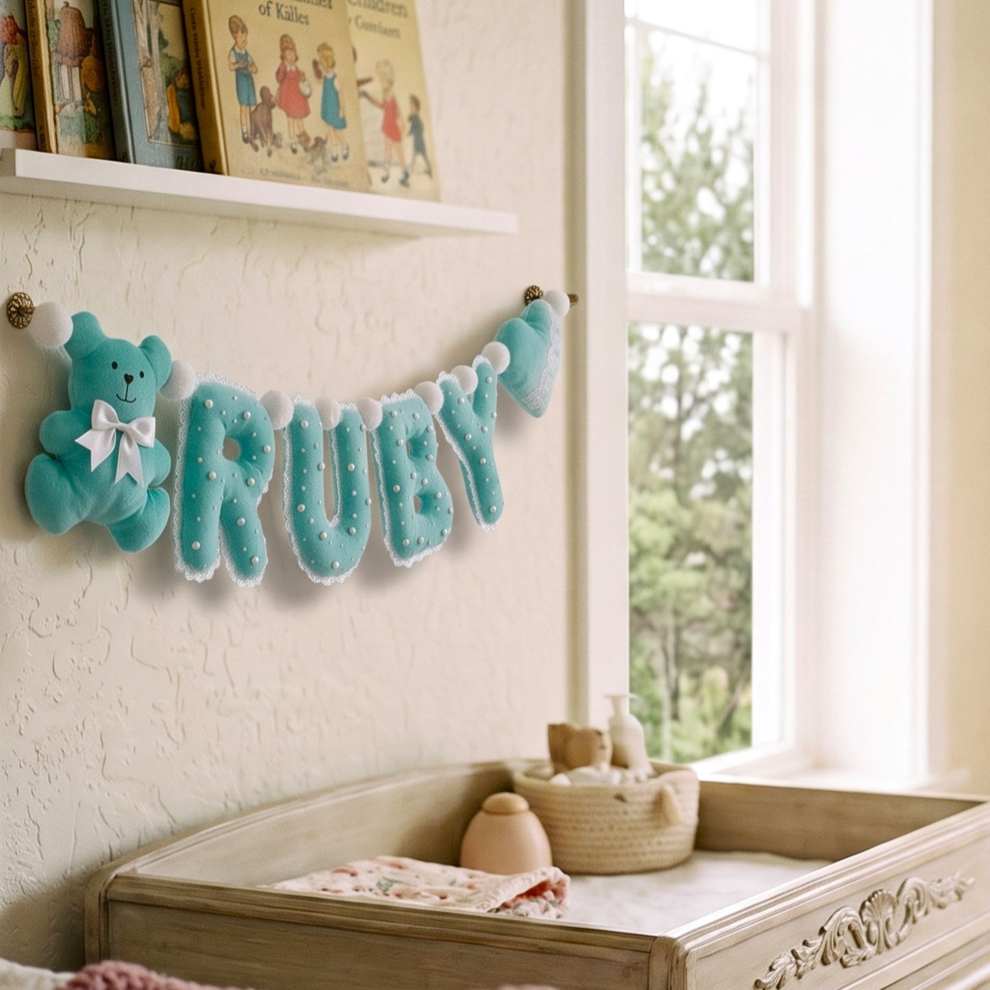 A light blue felt baby name banner spelling 'RUBY' with a matching plush teddy bear and bow, hanging on a nursery wall above a changing table near a bright window.