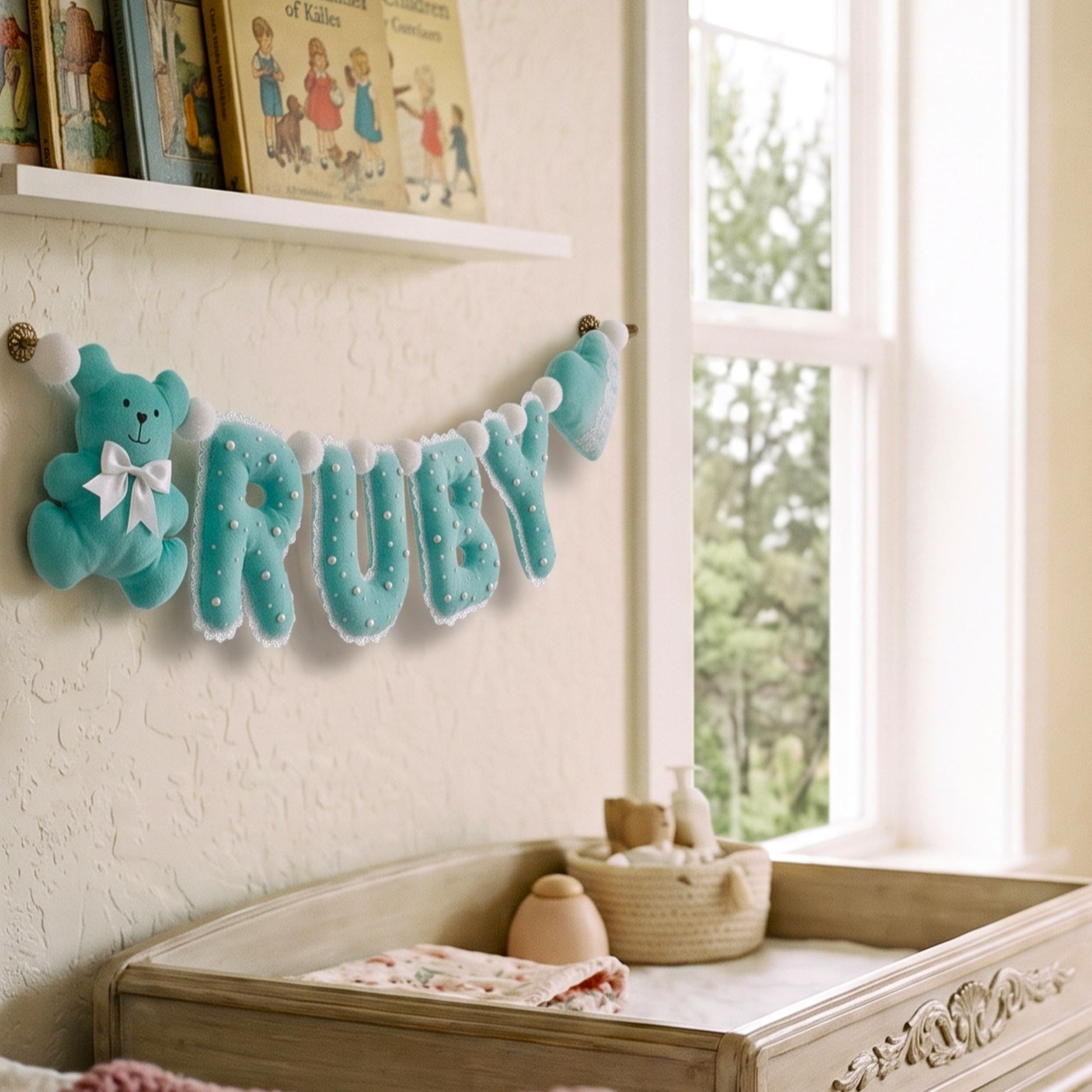 A light blue felt baby name banner spelling 'RUBY' with a matching plush teddy bear and bow, hanging on a nursery wall above a changing table near a bright window.