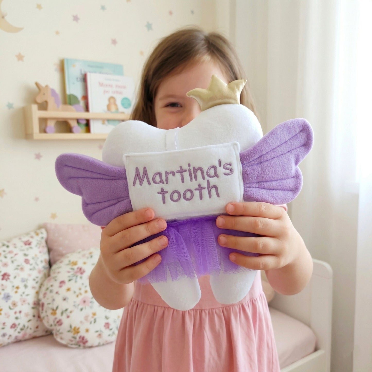 Child holding a purple tooth fairy pillow with 'Martina's tooth' embroidered on it in a room setting.
