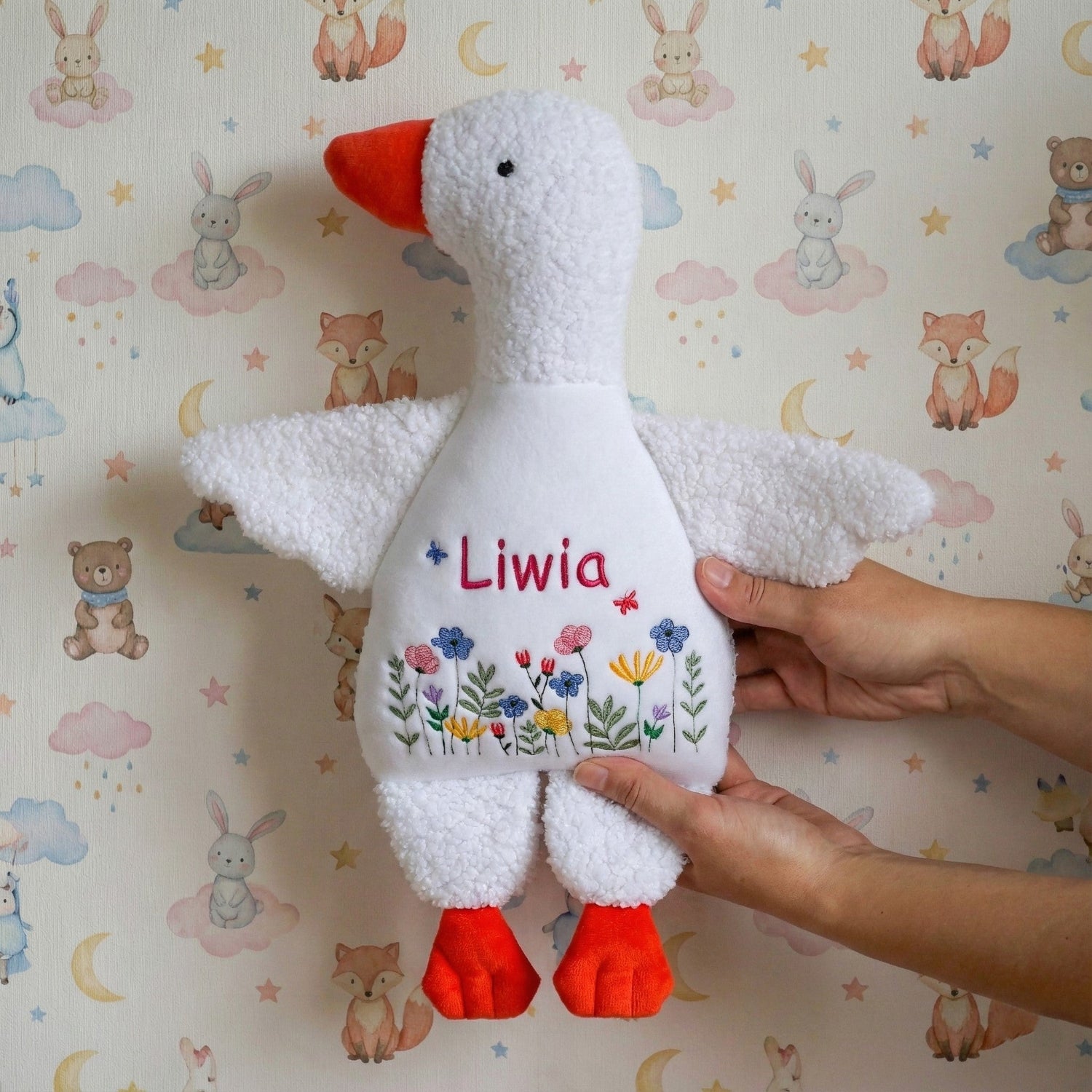 Plush toy goose with 'Liwia' embroidered on it, held against a wallpapered wall with animal illustrations.