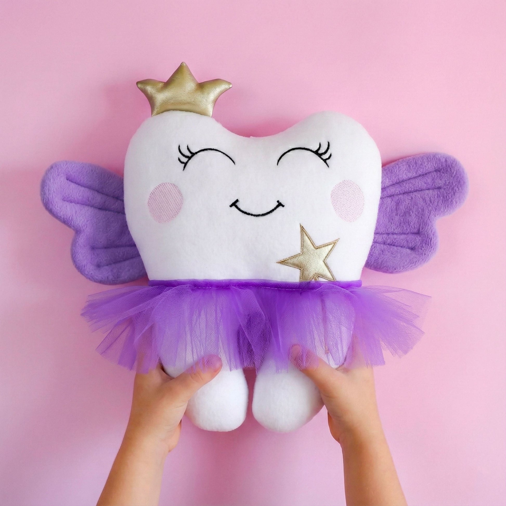 Plush tooth-shaped toy with fairy wings and a crown on a pink background