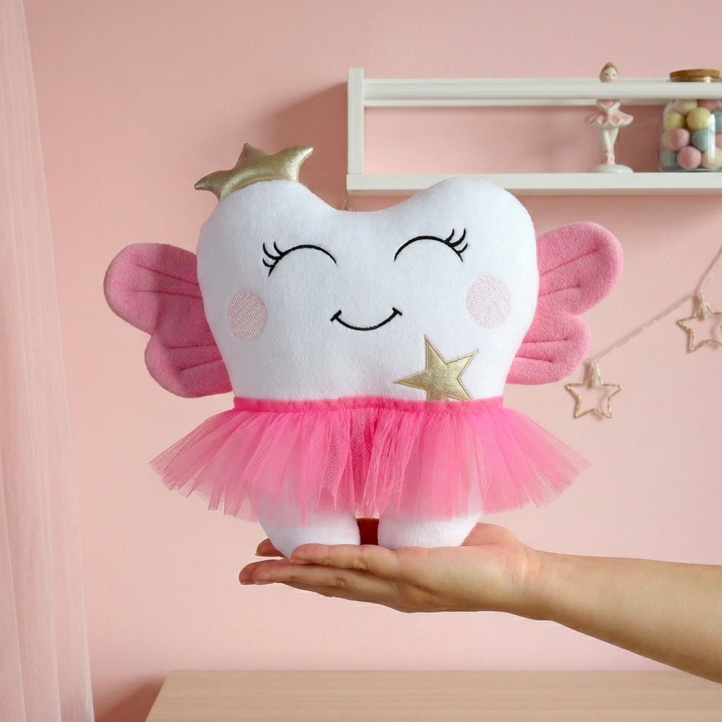 Plush tooth-shaped toy with fairy wings held by a hand against a pink background