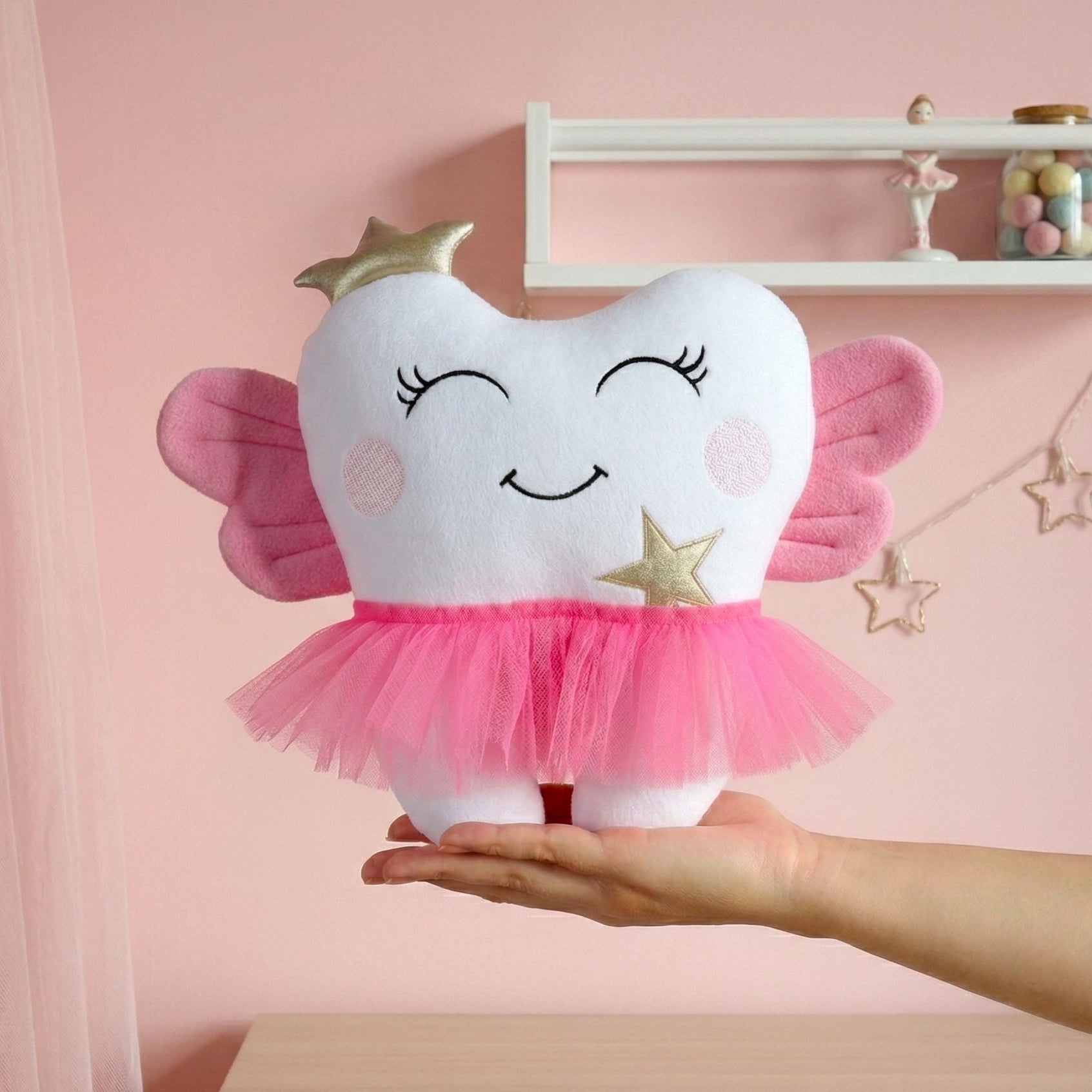 Plush tooth-shaped toy with fairy wings held by a hand against a pink background
