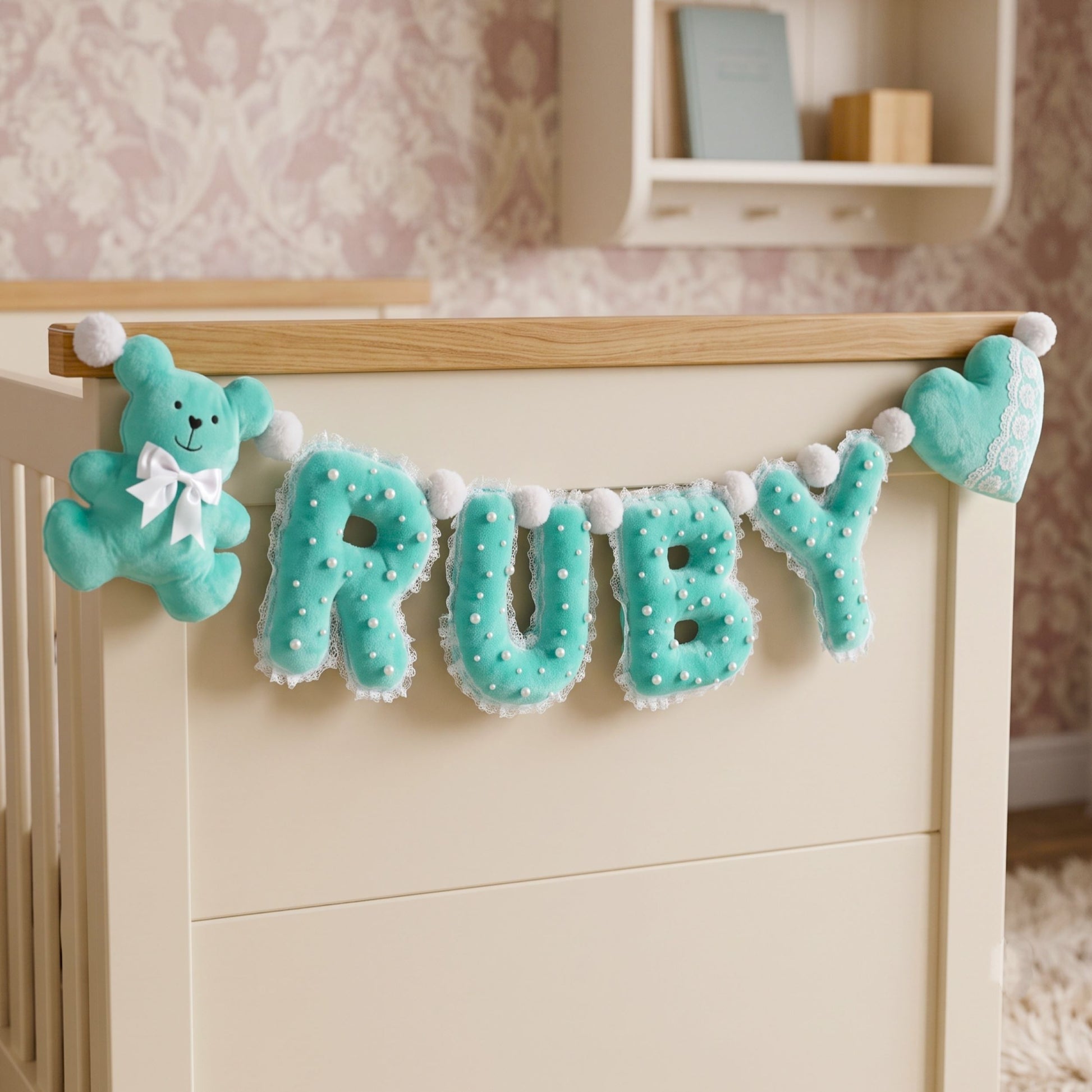 A personalized nursery banner featuring the name 'RUBY' in large turquoise letters with white pom-pom trim, hanging on a wooden crib bar. The banner includes a matching turquoise teddy bear plush on the left with a white bow and a heart-shaped plush on the right, set against a soft pink patterned nursery background.