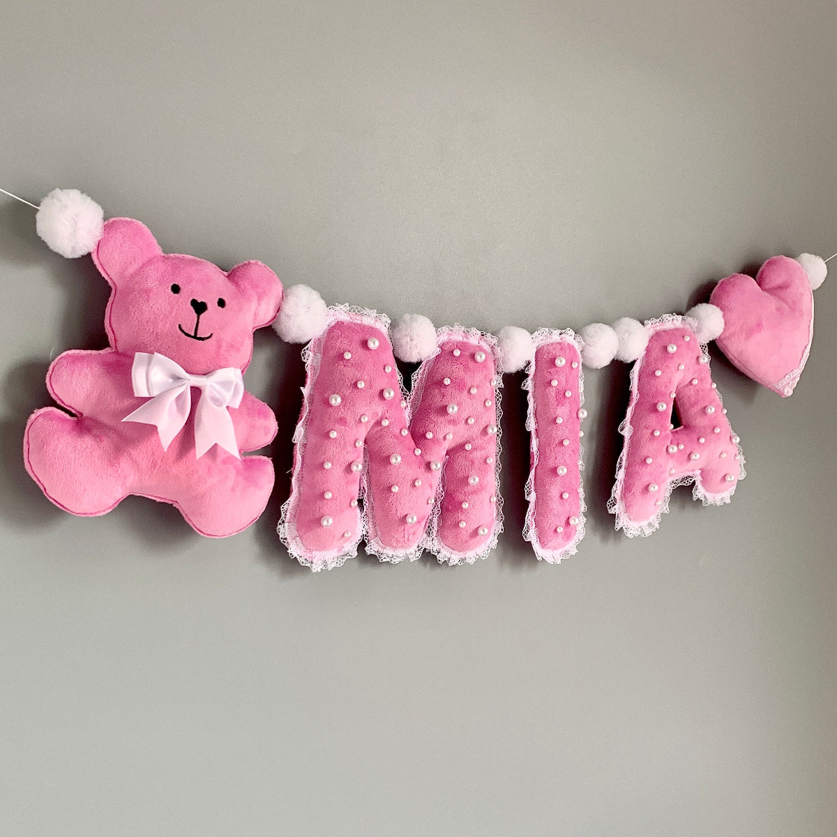 A pink personalized nursery wall banner featuring a plush teddy bear on the left, the name 'MIA' in decorative letters in the center, and a heart on the right, all hanging against a plain gray wall.