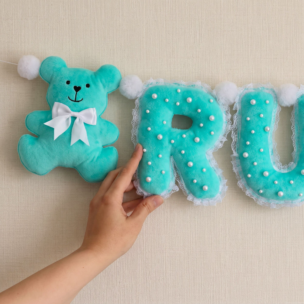 A hand holds a turquoise letter 'U' plush letter banner with white pom-poms and lace trim next to a matching plush teddy bear on a beige textured background.