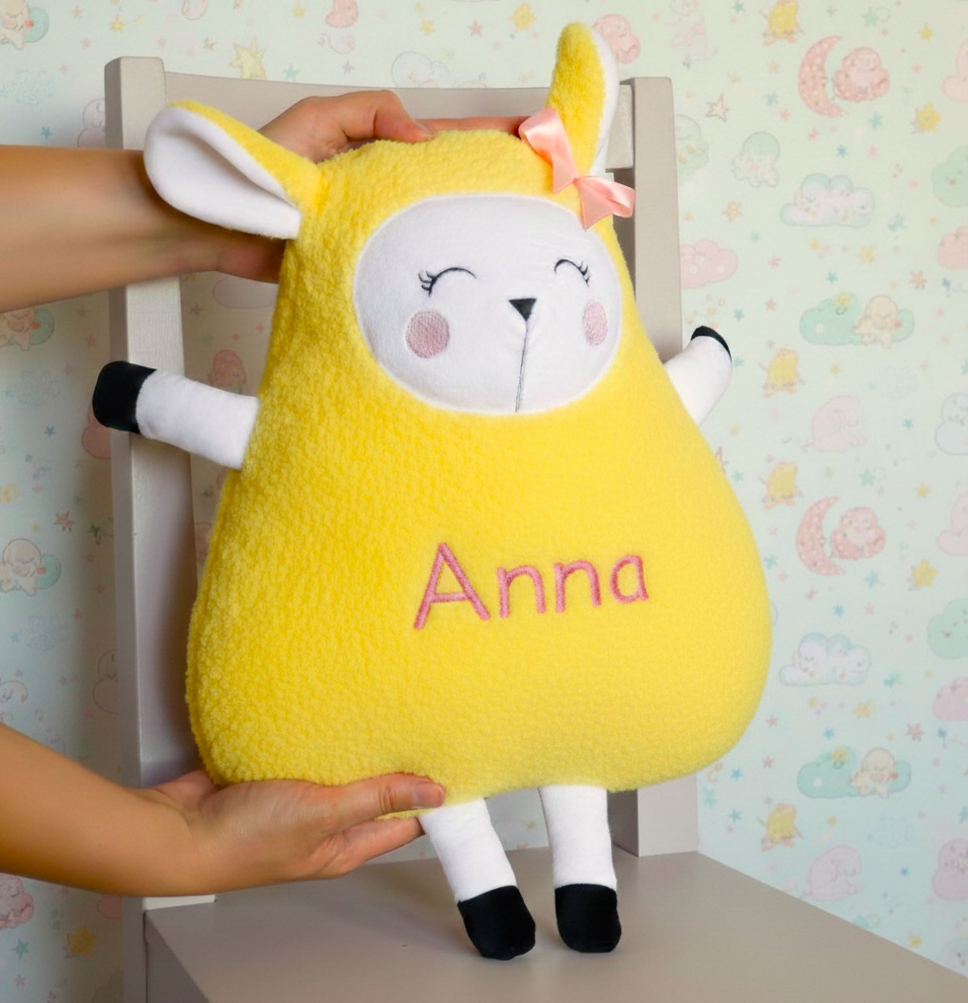 Yellow plush toy with 'Anna' embroidered on it, held by a person against a pastel wallpapered wall.