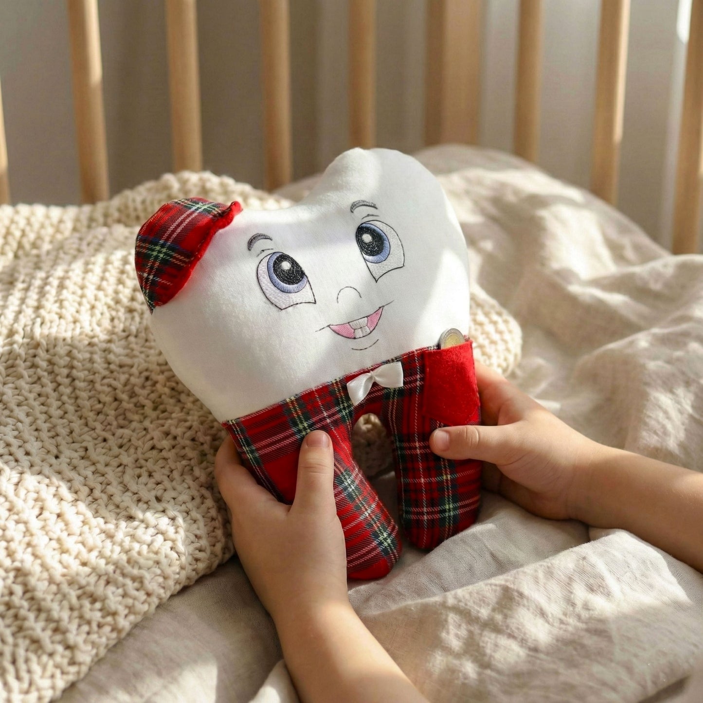 Plush tooth-shaped toy with a face and plaid outfit held by a child's hands on a bed.